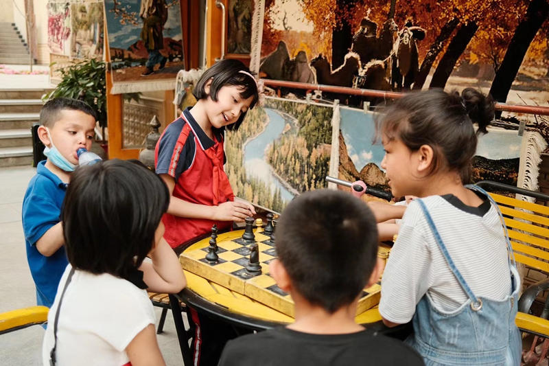 Dos niñas Uygur juegan al ajedrez juntos después de la escuela mientras sus amigos miran el juego frente a una tienda de pintura al óleo en Tuancheng, la ruta peatonal más famosa de la ciudad de Hotan, en la región autónoma Uygur de Xinjiang, en el noroeste de China, 22 de mayo de 2021. (Pueblo en Línea / Zhang Ruohan)