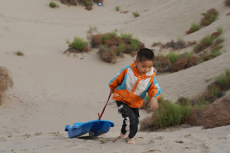 Preparándose para deslizarse por una pendiente de arena, un niño arrastra su trineo hasta la cima de la duna en la sección suroeste del desierto de Taklamakan en la región autónoma Uygur de Xinjiang, noroeste de China, el 21 de mayo de 2021 (Pueblo en Línea / Zhang Ruohan)