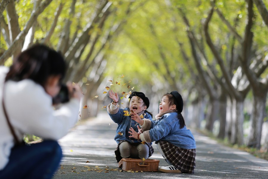 Una mujer toma fotos de niñas que juegan con hojas caídas en un parque del distrito de Jiangdu, en Yangzhou, provincia oriental china de Jiangsu, el 18 de noviembre de 2023. (Xinhua/Meng Delong) 