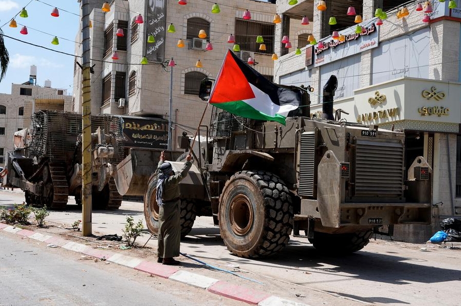 Un hombre palestino sostiene una bandera nacional frente a vehículos militares israelíes durante una incursión israelí en el campo de refugiados de Tulkarm, en el norte de Cisjordania, el 22 de agosto de 2024. (Xinhua/Ayman Nobani)